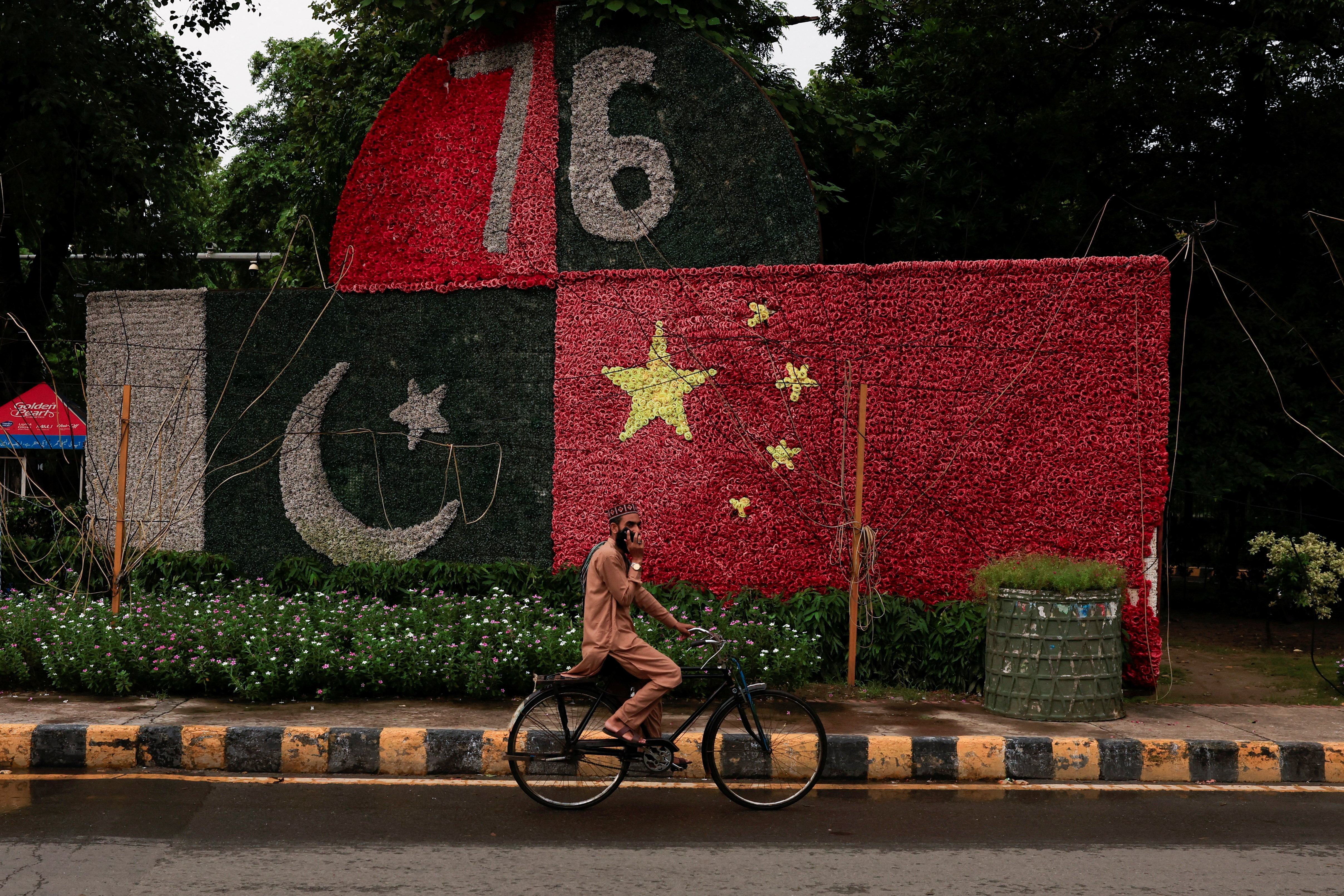 A man uses a mobile phone as he rides on a bicycle past a decorated wall depicting Pakistan and China's flags along a road.