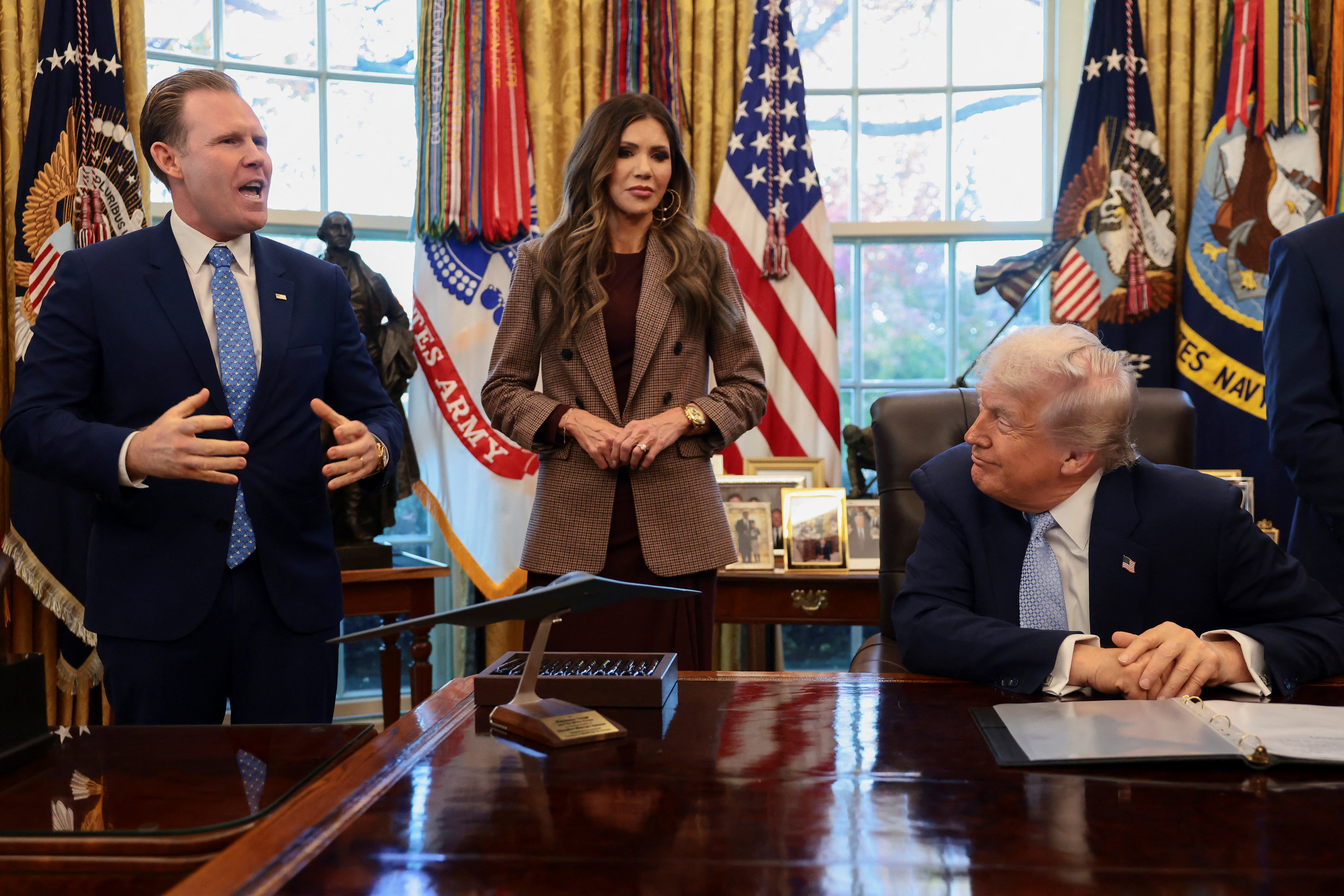 US President Donald Trump sits while Andrew Giuliani, head of the White House Task Force for the FIFA World Cup 2026, and US Homeland Security Secretary Kristi Noem stand by his side.