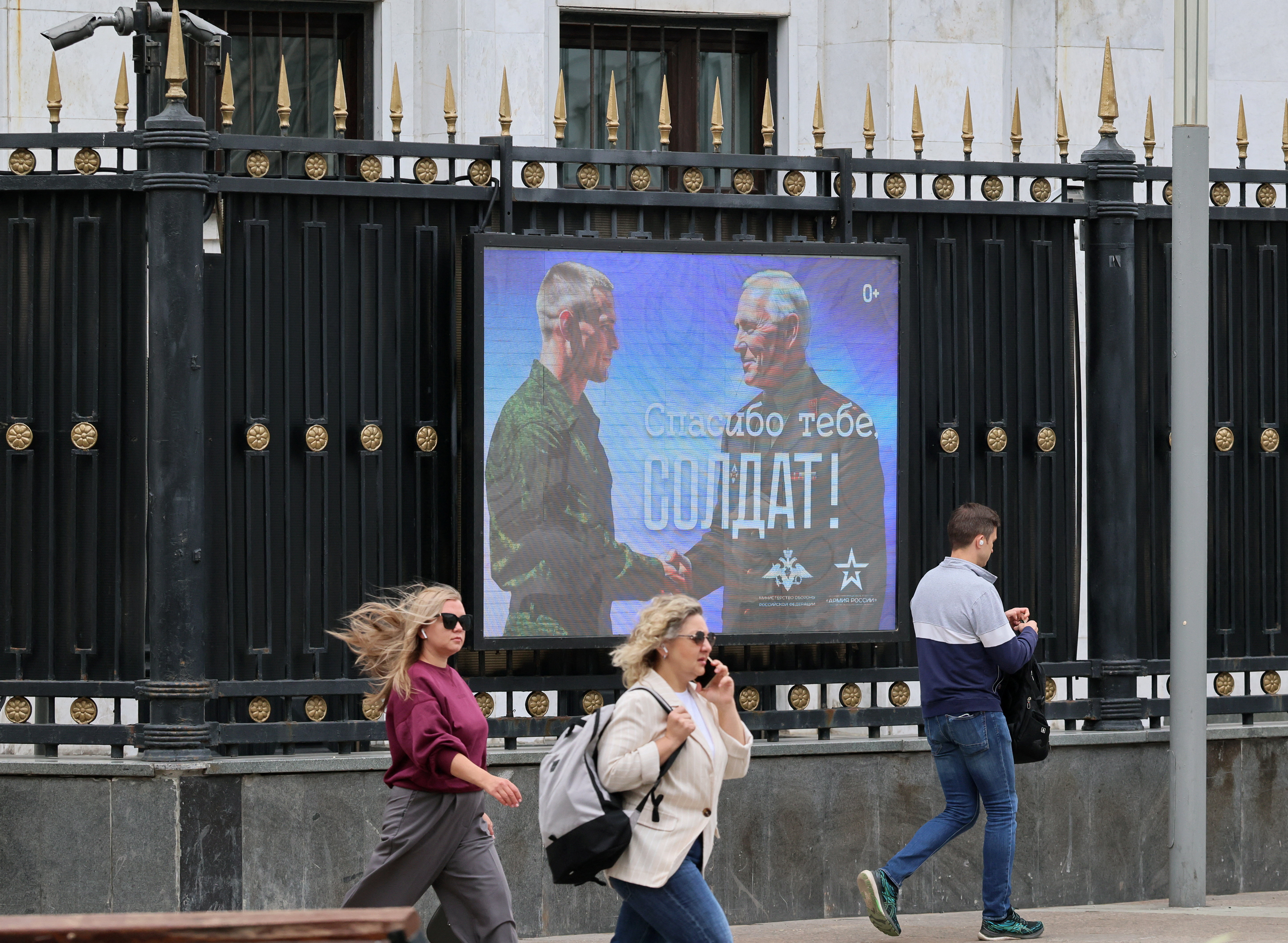 People walk past a screen that displays the message 'Thank you, soldier!', in Russian, with two men shaking hands.