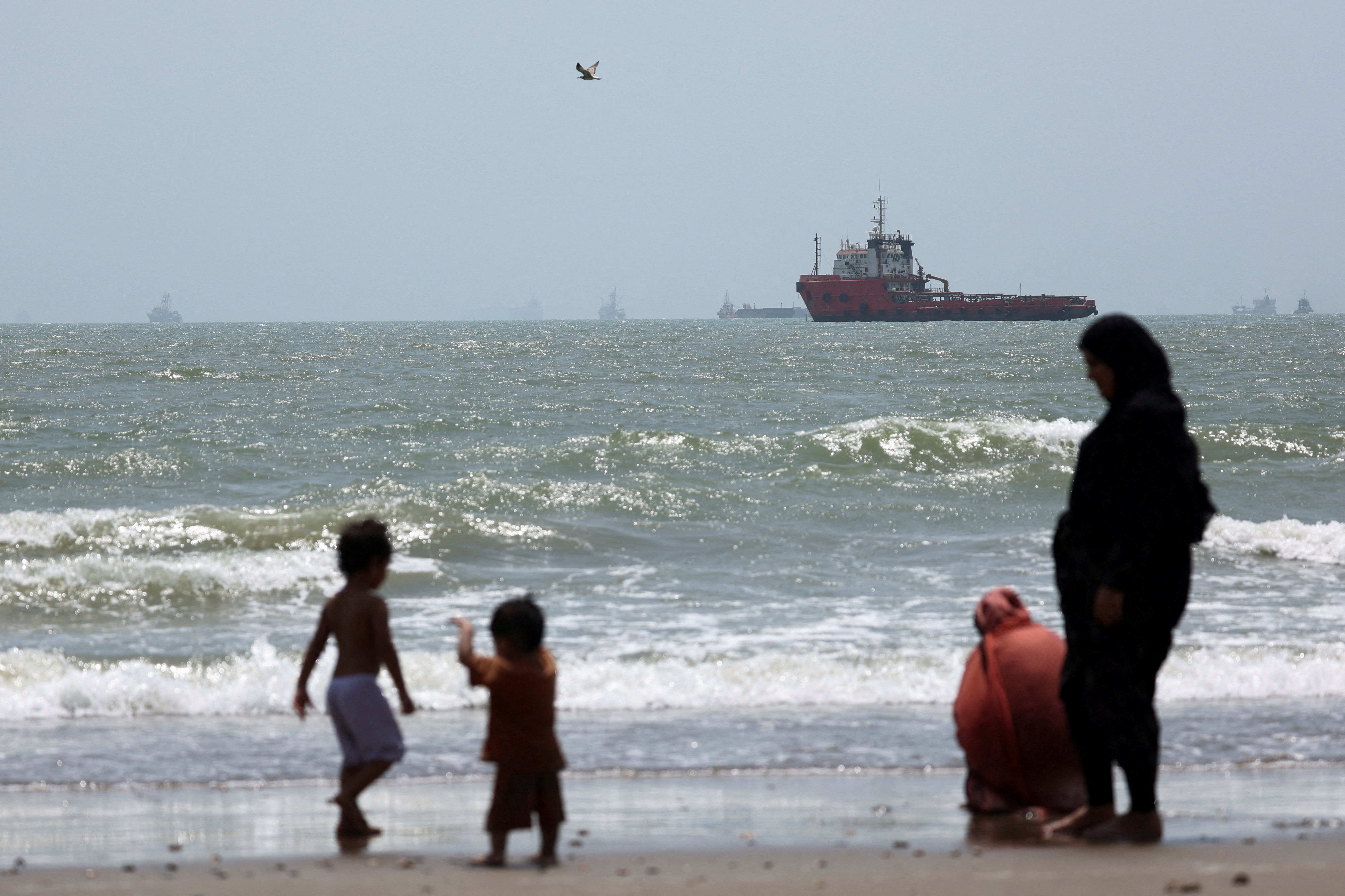 People gather on the beach, as vessels in the Strait of Hormuz are visible near the beach of Bandar Abbas, Iran, August 25, 2026. [Majid Asgaripour/WANA via Reuters]