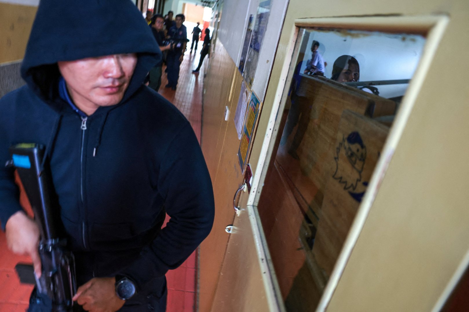 A police officer acting as an active shooter holds a rifle and walks past a classroom as students hide during a drill to prepare for an active shooter scenario at Surasakmontree School in Bangkok, Thailand on Aug. 19, 2026, following a deadly shooting at Debsirin Nonthaburi School in Nonthaburi province. [Athit Perawongmetha/Reuters]