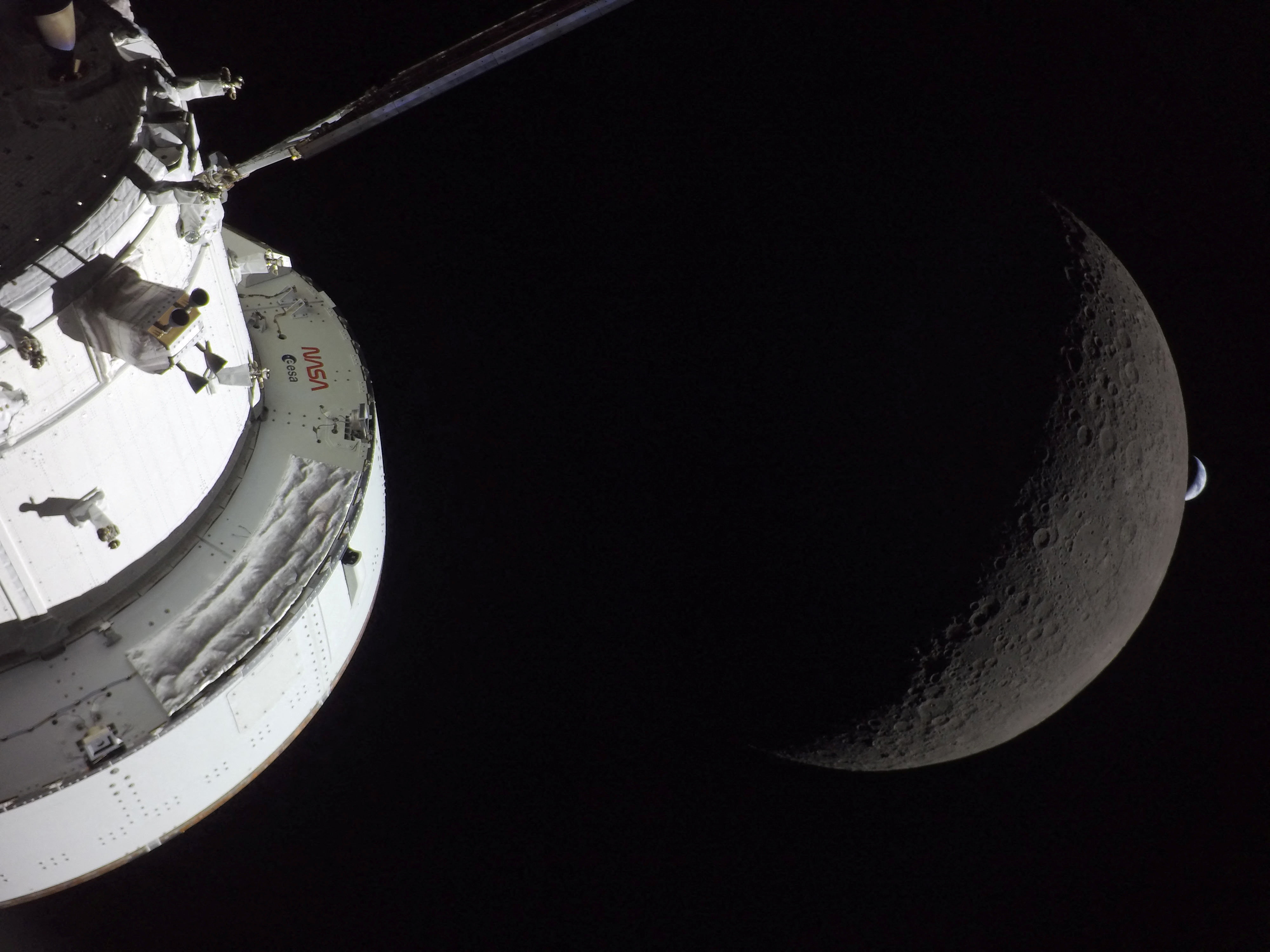 A view of NASA’s Orion spacecraft in the foreground, lit up by the sun as the moon and Earth are visible behind it as photographed by one of the cameras on the Orion spacecraft’s solar arrays during the Artemis II crew’s flyby of the moon on April 6, 2026