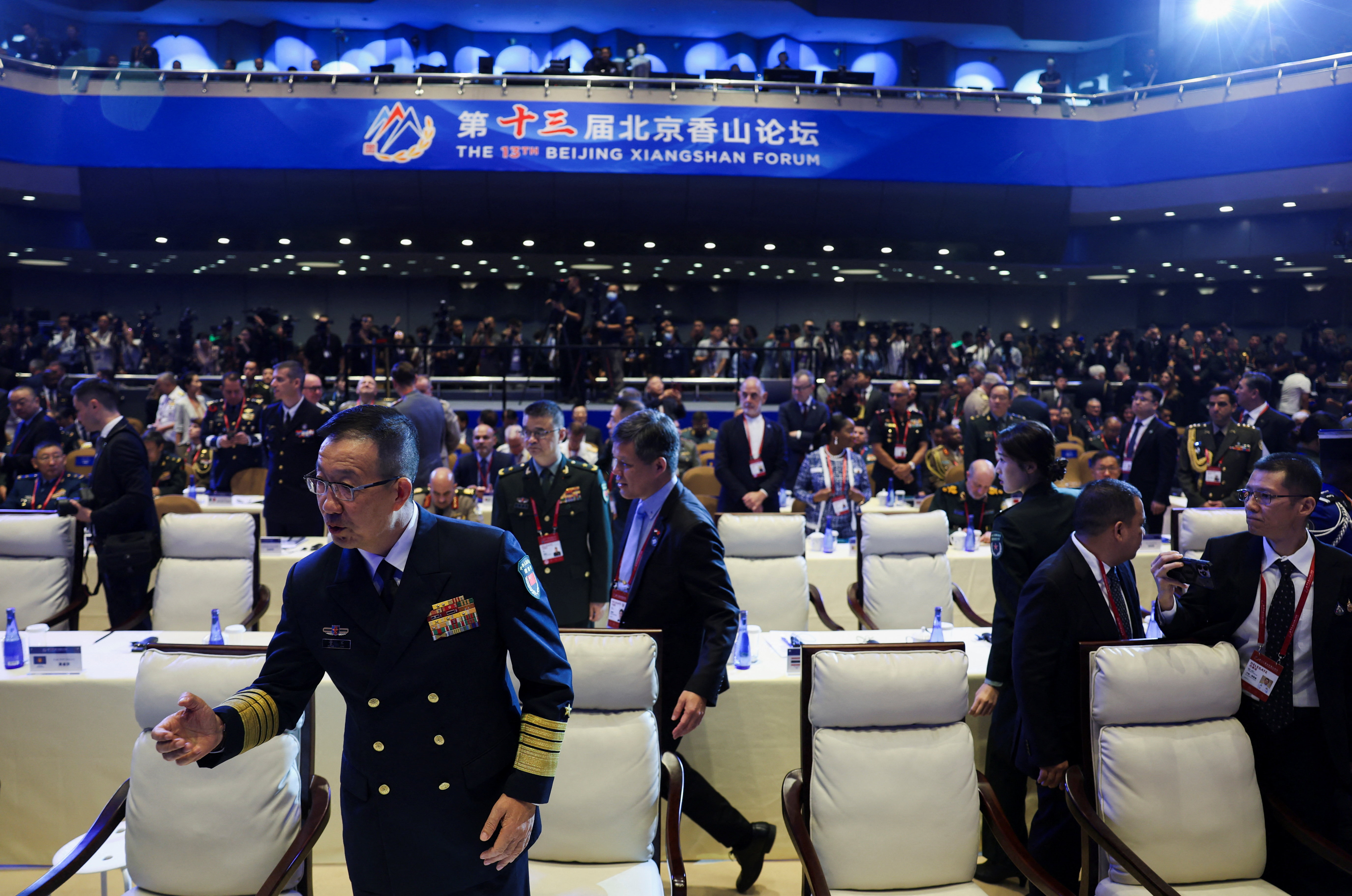 Men in dark suits and military uniforms walk together through a conference hall entrance.