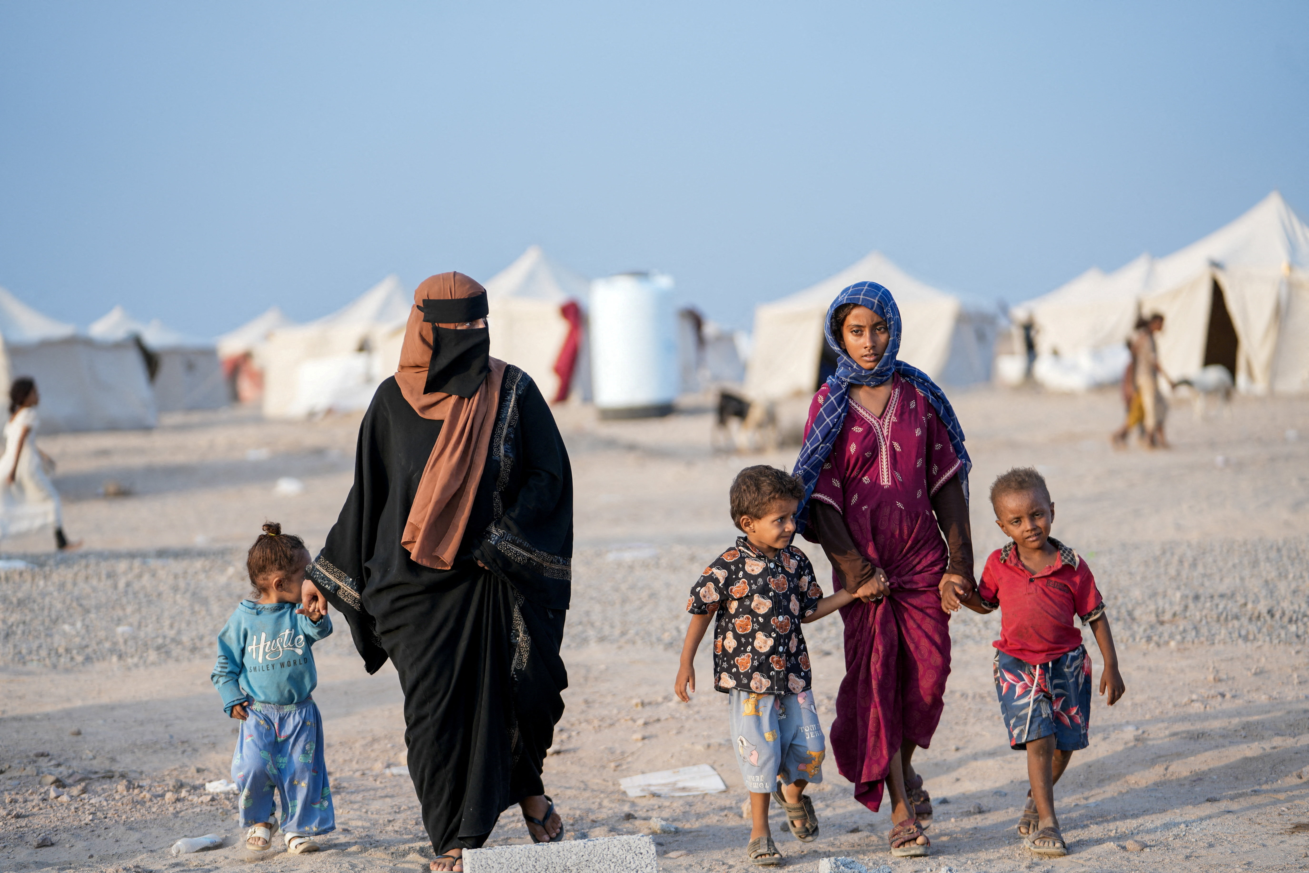 Women in black abayas and several children walking between tents at a displacement camp.