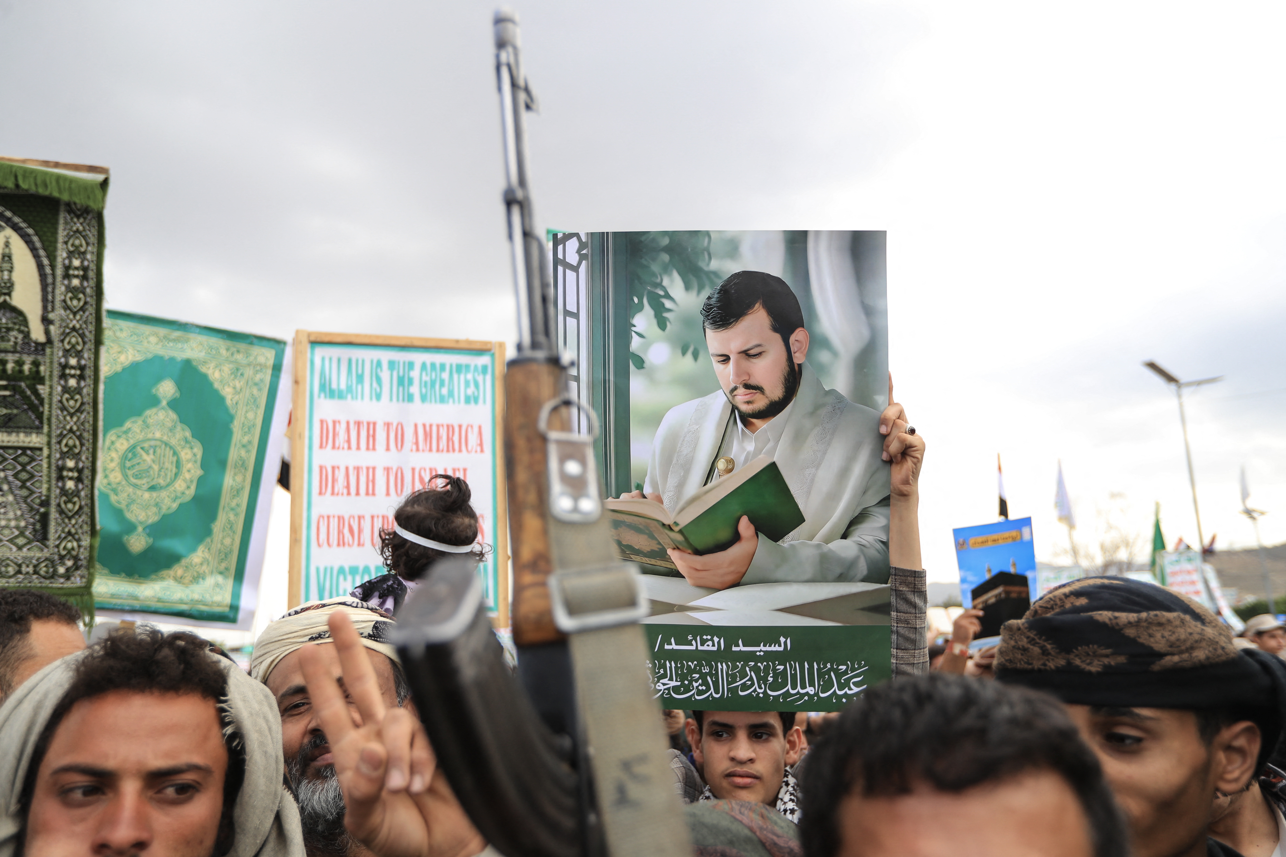 A dense crowd of men fills a street, many raising rifles above their heads, while one holds up a large printed portrait of a bearded man in a turban.