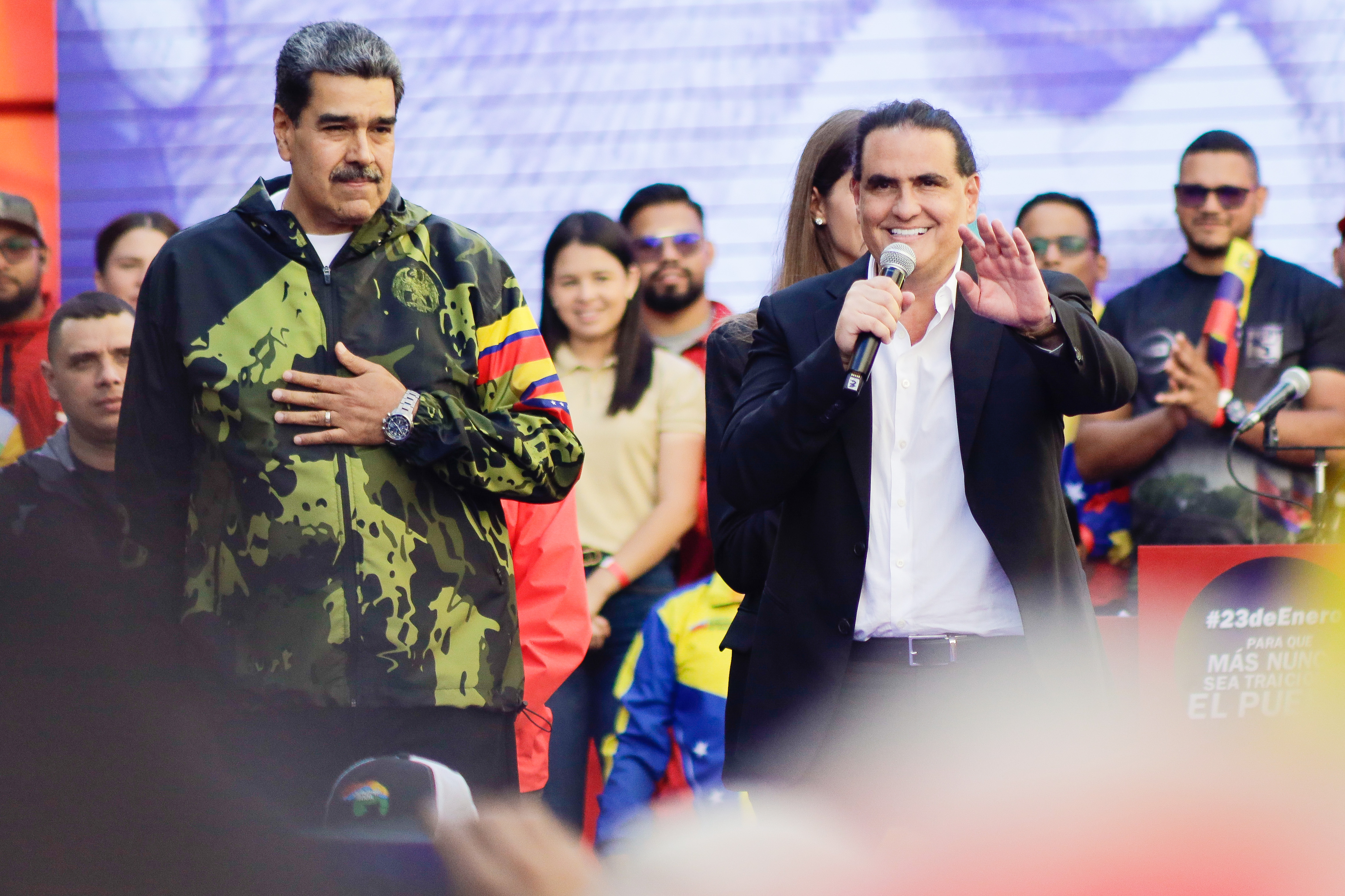 Two men in dark jackets stand together at a microphone in front of a crowd.