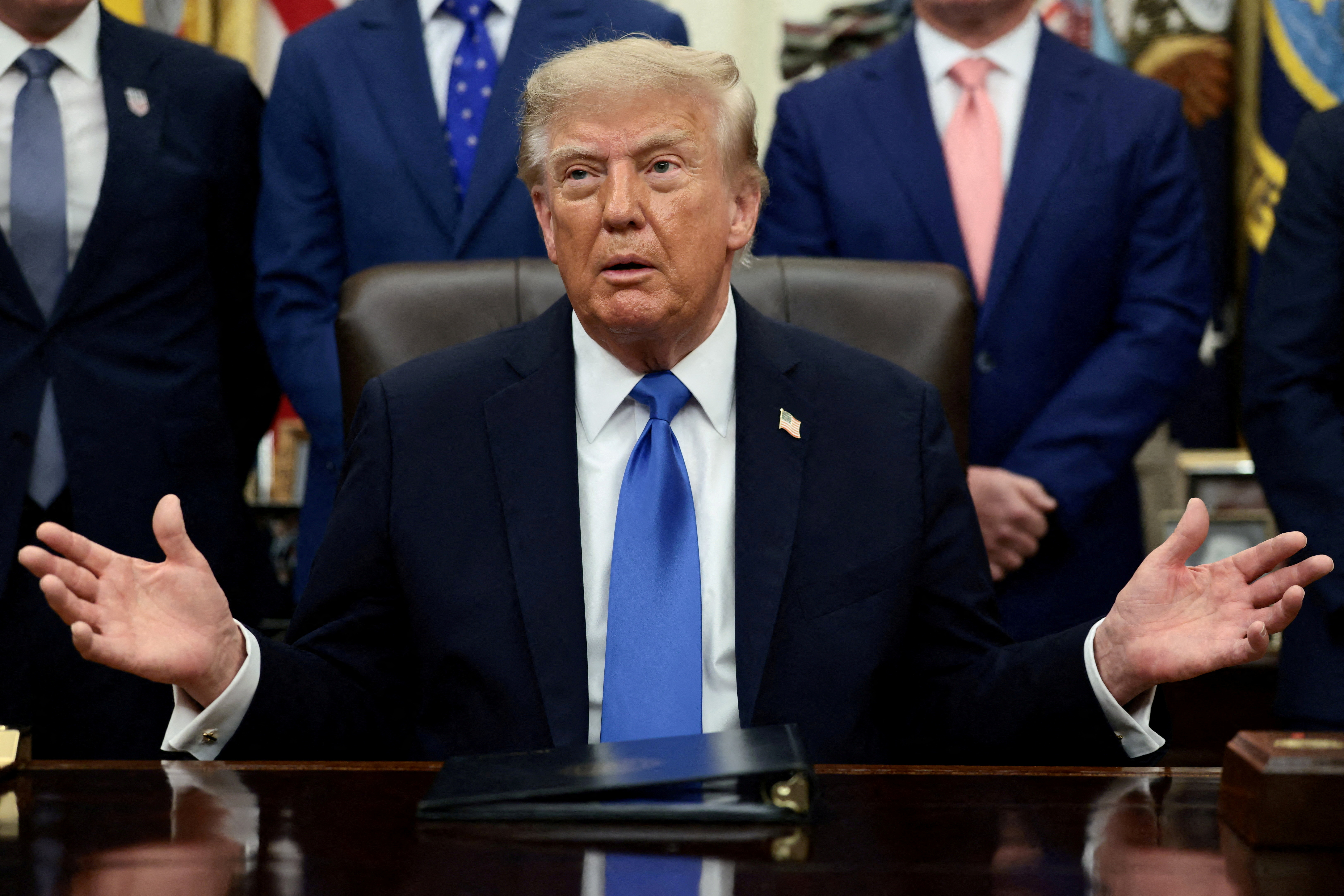 A man in a dark suit speaks while seated behind a desk in a formal office.