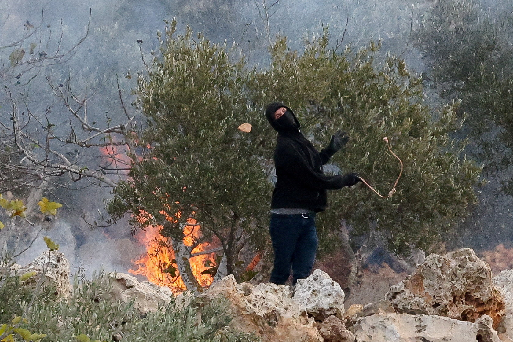 An Israeli settler casts a stone towards Palestinians, next to a fire, amid clashes during Palestinians' olive harvest in Abu Falah near Ramallah in the occupied West Bank on October 16, 2026. [Ammar Awad/Reuters]